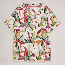 Load image into Gallery viewer, A short-sleeve V-neck scrub top with a colorful tropical floral print on an off-white background, laid flat on a light gray textured surface.
