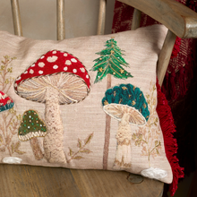 Load image into Gallery viewer, A square decorative throw pillow with embroidered woodland mushrooms and a pine tree on a beige linen background, resting on a wooden chair.