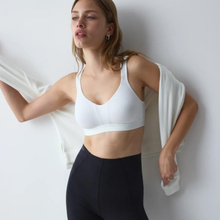 Load image into Gallery viewer, A woman wearing a white textured sports bra and black leggings, holding a white cardigan against a plain light gray wall.