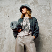 Load image into Gallery viewer, A woman posing against a textured concrete wall wearing a gray fuzzy bucket hat, a dark gray knit cardigan over a gray graphic t-shirt, and light gray wide-leg trousers.