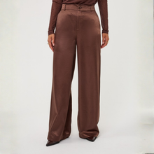 Load image into Gallery viewer, A model is shown from the waist down wearing high-waisted, wide-leg brown trousers with a subtle satin sheen, paired with a matching brown long-sleeve top and pointed-toe heels, against a plain white studio background.