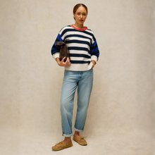 Load image into Gallery viewer, A woman stands against a textured beige studio backdrop wearing a navy and white striped knit sweater with a red collar, light blue jeans, tan loafers, and holding a dark brown clutch.