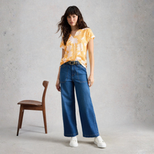 Load image into Gallery viewer, Woman wearing a yellow tie-dye shirt and blue jeans standing against a gray background with a wooden chair.