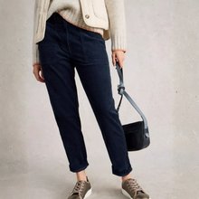 Load image into Gallery viewer, A person wearing dark navy blue jeans, a cream cardigan, and brown sneakers, holding a black shoulder bag against a textured light gray wall.