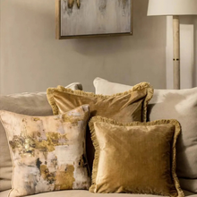 Load image into Gallery viewer, Three decorative throw pillows arranged on a beige sofa in a styled living room setting. One pillow features an abstract gold, beige, and white textured pattern, while the other two are solid golden-brown velvet with fringed edges.