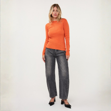 Load image into Gallery viewer, A woman modeling a bright orange long-sleeve knit sweater paired with wide-leg faded gray jeans and black pointed-toe heels, standing against a plain off-white studio background.
