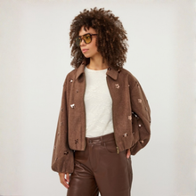 Load image into Gallery viewer, A model with curly brown hair and sunglasses wearing a cropped brown suede jacket with embroidered flower details, a white textured top, and brown faux-leather pants, set against a plain light gray background.
