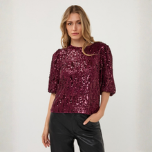 Load image into Gallery viewer, A woman modeling a burgundy sequined short-sleeve top with puffed sleeves and black pants, standing against a plain light gray studio background.
