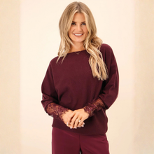 Load image into Gallery viewer, A smiling blonde woman wearing a burgundy long-sleeve knit top with lace sleeves and matching burgundy pants, standing against a soft beige background.

