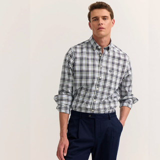 A man wearing a long-sleeve button-down shirt with a Green plaid pattern, paired with navy blue trousers, standing against a plain off-white background.