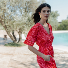 Load image into Gallery viewer, Woman in a red dress standing by a pool with trees in the background
