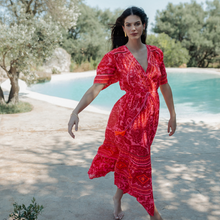 Load image into Gallery viewer, Woman in a red dress standing by a pool with trees in the background
