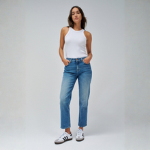 Load image into Gallery viewer, A woman modeling a pair of straight-leg blue jeans with a white sleeveless top and white sneakers, standing against a light gray studio background.
