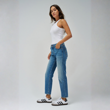Load image into Gallery viewer, A woman modeling a pair of high-waisted, straight-leg blue jeans, paired with a white tank top and white sneakers with black stripes, standing against a light gray studio background.
