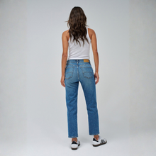 Load image into Gallery viewer, A woman is shown from behind wearing a pair of medium-wash blue jeans, a white tank top, and white sneakers with black stripes, standing against a light gray studio background.
