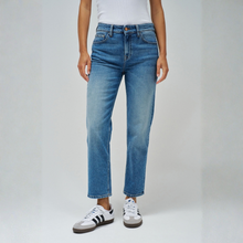 Load image into Gallery viewer, A model wearing a pair of medium-wash blue straight-leg jeans, styled with a white t-shirt and white sneakers with black stripes, against a light gray studio background.
