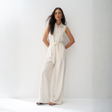 Load image into Gallery viewer, A woman stands against a plain white studio wall wearing a sleeveless cream-colored jumpsuit with thin vertical stripes, a button-up front, and a matching tie belt at the waist.
