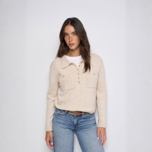 Load image into Gallery viewer, A woman modeling a light beige knit polo sweater with a buttoned collar and chest pockets, worn over a white t-shirt and blue jeans, against a plain light gray background.
