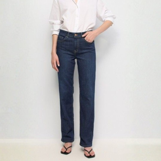 A model wearing dark blue straight-leg jeans, a white button-down shirt, and black sandals, standing against a plain off-white wall.