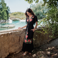 Load image into Gallery viewer, Woman in a black dress with pink floral embroidery standing by a poolside.
