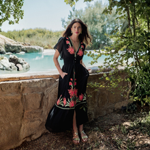 Load image into Gallery viewer, Woman in a black dress with floral embroidery standing by a poolside.
