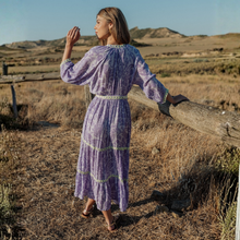 Load image into Gallery viewer, Woman in a purple dress standing in a field with wooden posts.
