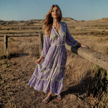 Load image into Gallery viewer, Woman in a long, patterned dress standing in a desert landscape with a wooden fence.
