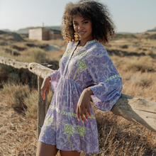 Load image into Gallery viewer, Woman in a purple dress with green patterns standing in a desert-like landscape.
