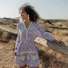 Load image into Gallery viewer, Woman in a purple dress with green patterns standing in a desert landscape.
