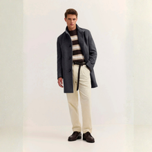 Load image into Gallery viewer, A male model stands in a studio wearing a dark charcoal gray overcoat over a beige and brown striped sweater, cream trousers, a black belt, and dark brown leather shoes.
