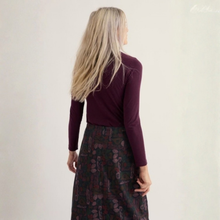 Load image into Gallery viewer, A woman with long blonde hair is shown from behind wearing a dark plum long-sleeve top and a dark patterned skirt against a plain off-white background.
