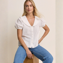 Load image into Gallery viewer, Woman wearing a white blouse with ruffled collar and blue jeans sitting on a wooden stool against a beige background.
