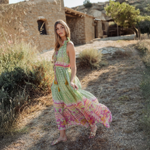 Load image into Gallery viewer, Woman in a colorful dress standing on a dirt path with rustic buildings in the background
