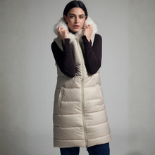 Load image into Gallery viewer, A woman wearing a long cream-colored puffer vest with a white faux-fur collar, layered over a dark brown long-sleeve top and blue jeans, set against a plain gray studio background.
