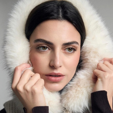 Load image into Gallery viewer, Close-up portrait of a woman wearing a white faux-fur hooded outerwear with dark ribbed knit sleeves, against a soft gray studio background.
