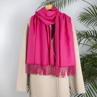 A bright pink scarf with fringe trim draped over a beige coat on a wooden hanger, set against a light gray wall with green palm leaves in the background.