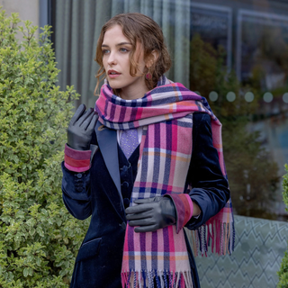 A woman outdoors wearing a thick pink, purple, and blue plaid scarf with fringe, black gloves, and a dark navy velvet coat.