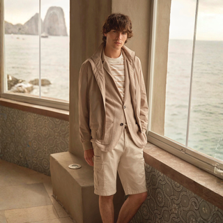Person wearing a beige jacket and shorts standing by large windows with a scenic view.