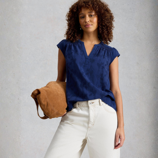 Woman wearing a blue top and white pants, holding a brown bag against a gray background