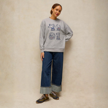 Load image into Gallery viewer, A woman modeling a light gray crewneck sweatshirt with a blue graphic print of four cartoon dog heads, paired with wide-leg cuffed jeans and brown sneakers, standing against a textured beige wall.
