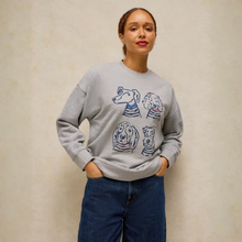 Load image into Gallery viewer, A woman wearing a light gray crewneck sweatshirt with a graphic print of four dogs wearing striped shirts, paired with dark blue jeans, standing against a textured beige wall.
