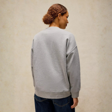 Load image into Gallery viewer, A woman viewed from behind wearing a light gray crewneck sweatshirt and dark blue jeans, standing against a textured beige wall.
