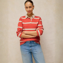 Load image into Gallery viewer, A woman wearing a red and white horizontally striped long-sleeve polo shirt with a light beige collar, paired with light blue jeans, standing against a neutral beige textured wall.
