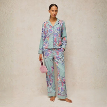 Load image into Gallery viewer, A woman stands barefoot against a textured beige wall wearing a matching pajama set in a light mint green color with a colorful floral print in pink, purple, and blue. She is holding a small pink fuzzy handbag.
