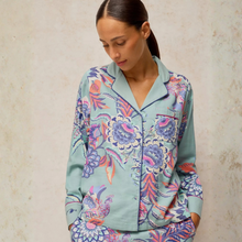 Load image into Gallery viewer, A woman wearing a light blue pajama set with a colorful floral and bird print, standing against a textured beige wall.
