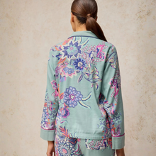 Load image into Gallery viewer, A woman shown from the back wearing a sage green satin pajama set with a vibrant multicolor floral print. The image features a textured beige background.
