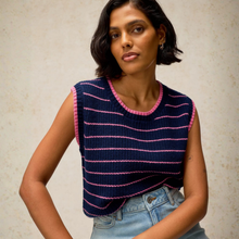 Load image into Gallery viewer, A woman wearing a sleeveless navy blue knit top with pink horizontal stripes and pink trim, paired with light-wash blue jeans, posing against a textured beige wall.
