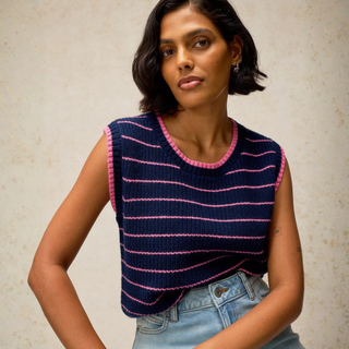 A woman wearing a sleeveless navy blue knit top with pink horizontal stripes and pink trim, paired with light-wash blue jeans, posing against a textured beige wall.