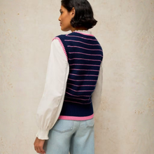 Load image into Gallery viewer, A woman shown from the back wearing a navy blue knit sweater vest with pink stripes and trim, layered over a white long-sleeve blouse and light blue jeans, set against a textured beige wall.
