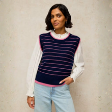 Load image into Gallery viewer, A woman wearing a navy blue ribbed knit sweater vest with pink trim over a white long-sleeve blouse and light blue jeans, standing against a textured beige wall.
